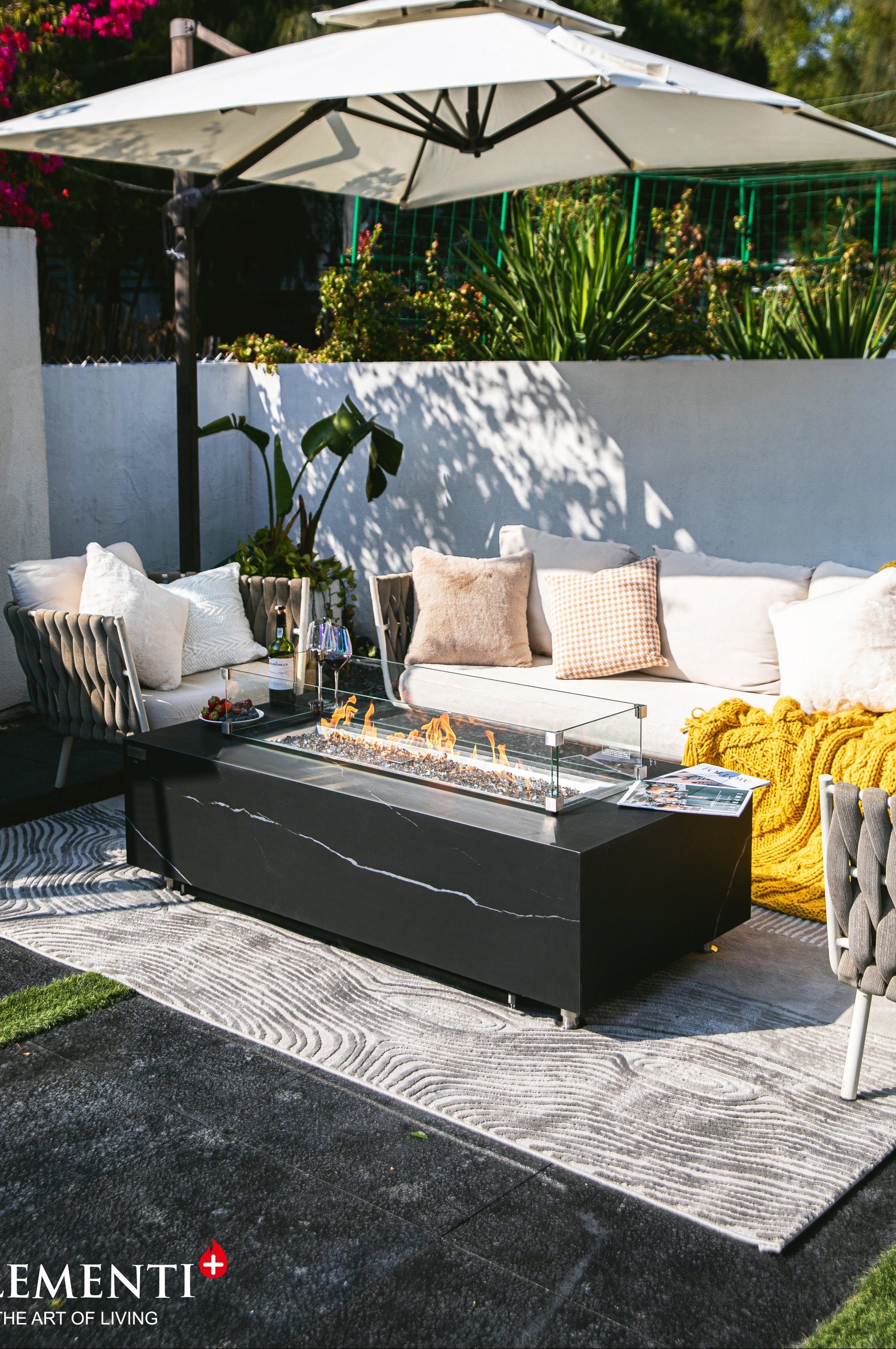 Outdoor patio furniture set featuring a black rectangular fire pit table with a glass wind guard, a white sectional sofa with neutral and yellow throw pillows, a gray patterned area rug, and a large white patio umbrella in a garden setting.